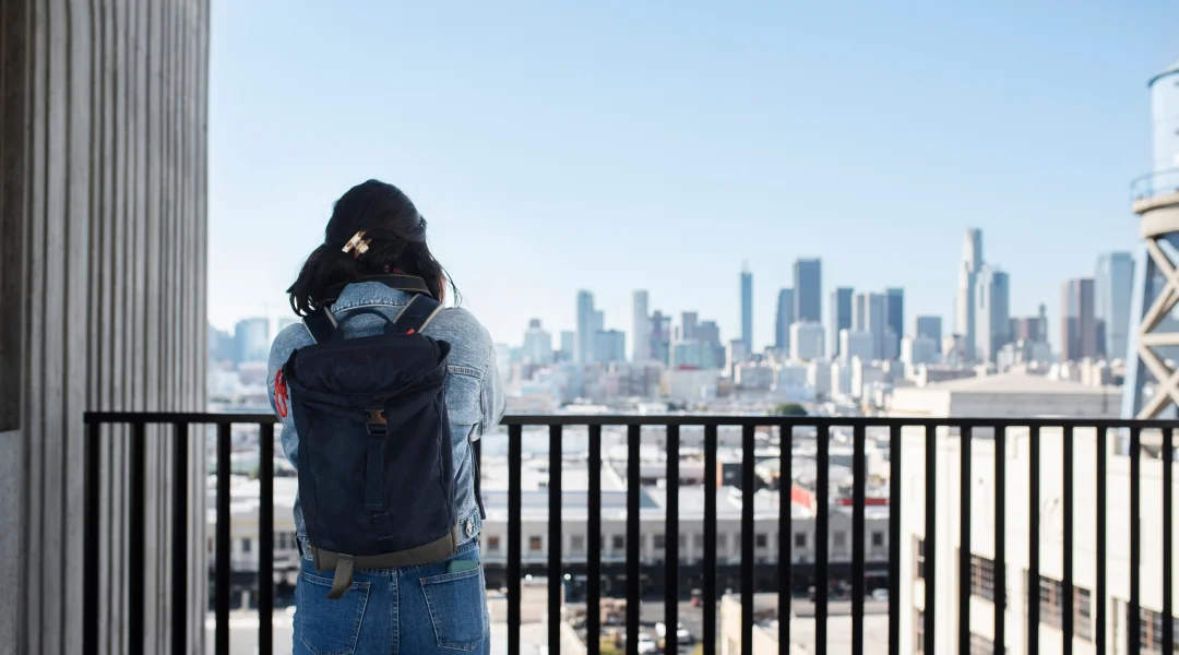A woman with a backpack stands on a balcony, gazing at a city skyline, symbolizing her new life in Canada.